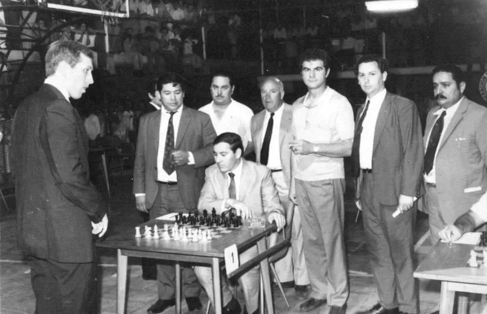 MOMENTO ESPECIAL. “Bobby” Fischer, parado frente al tablero, con Iván Rodríguez a punto de hacer una jugada. Parado, de remera clara y mirando a la cámara, Rubén Zusman. 