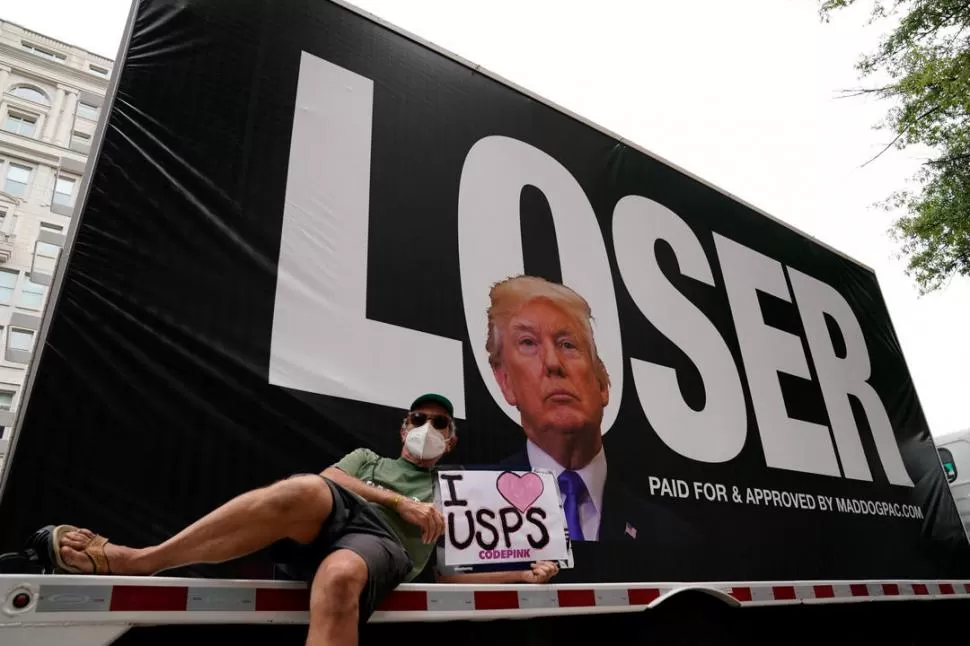 CAMPAÑA. Un hombre con barbijo muestra un cartel a favor del Servicio Postal de Estados Unidos, frente a un cartel que muestra a Trump como “perdedor”. REUTERS