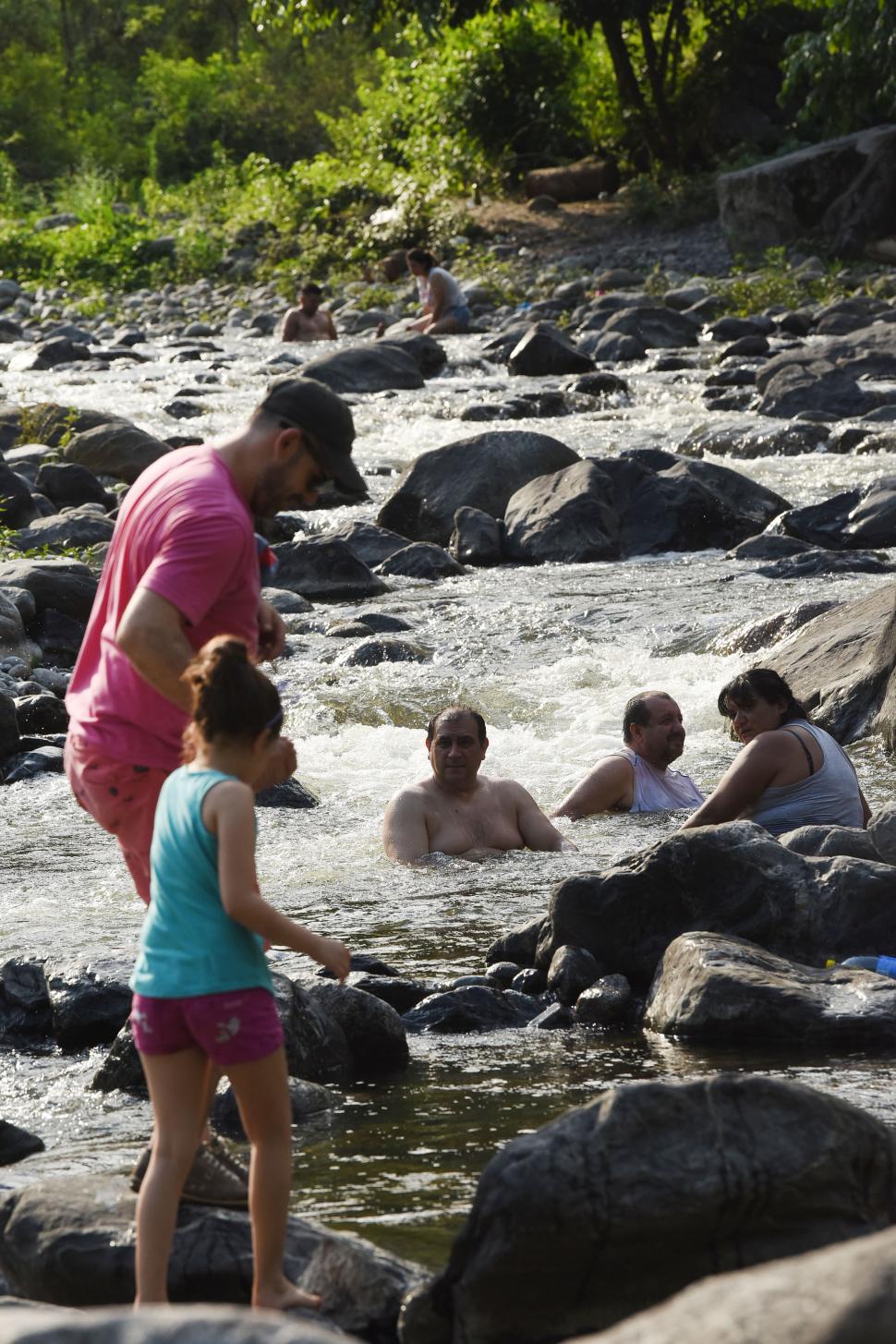 EN FAMILIA. Un padre ayuda a su hija a caminar entre las piedras y la corriente del río. Detrás, tres adultos los observan y enfrían sus cuerpos sumergidos en el agua.