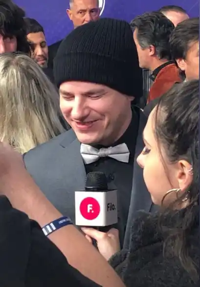 NAHUEL PENNISI. Traje y pajarita gris sobre camisa negra, más gorra de lana cubriéndole la cabeza, recientemente rapada.   