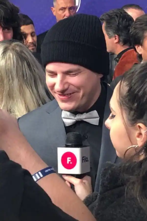 NAHUEL PENNISI. Traje y pajarita gris sobre camisa negra, más gorra de lana cubriéndole la cabeza, recientemente rapada.   