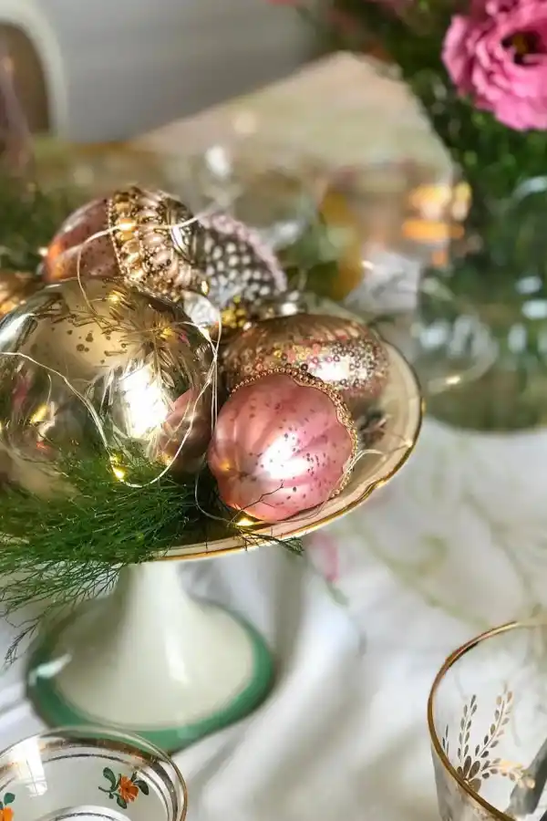 Pie de torta antiguo con adornos navideños vintage, lucecitas a pila y algunas ramas verdes de pino es una buena idea para el centro de tu mesa.