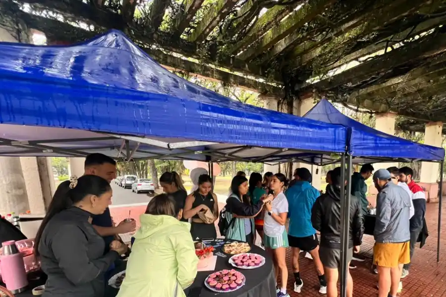 Bajo el gazebo, la lluvia se volvió apenas un murmullo. Sobre los tablones improvisados como mesas, se desplegaban tortas caseras, alfajores de maicena, frutas cortadas y termos con mate y café.